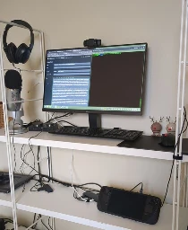 image of a steam deck, USB-C dock, external monitor, and cable spaghetti - all squished onto a cluttered and unstable bookshelf, in lieu of a desk. 