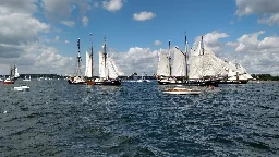 Photo of two three-mast sailing ships with full sails on a sunny day, surrounded by several smaller boats.